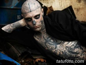 Фото тату: мужчина с татуировкой черепа на лице и шее, сидящий на фоне металлолома и мусора.
